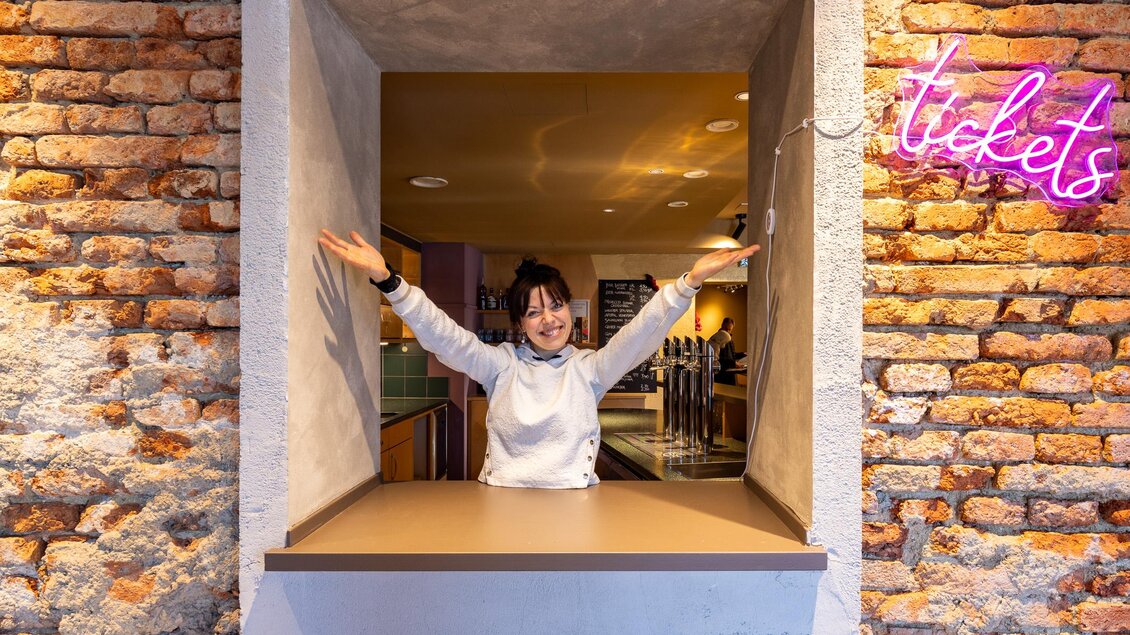 Jula Zangger steht mit erhobenen Armen am Ticketschalter im Café Marie vor einer beleuchteten Backsteinwand. | © Harry Schiffer Photodesign