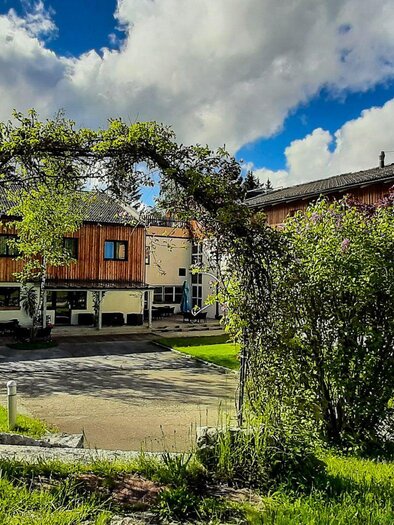 <p>Die Gebäude des JUFA Hotels mit Holzverkleidung und großen Fenstern.Im grünen Garten gibt es hohe Sträuche und einen Bogen. Im Hintergrund befindet sich eine Terrasse mit Tischen und Sonnenschirmen.</p> | © JUFA Hotels