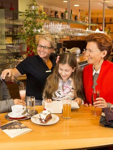 Eine gemütliche Kaffeepause in einem Café mit einer Familie und einer freundlichen Kellnerin. Sie genießen Getränke und einen Kuchen in einer einladenden Atmosphäre. | © Wolfgang Hummer - Kaufhaus Hubmann