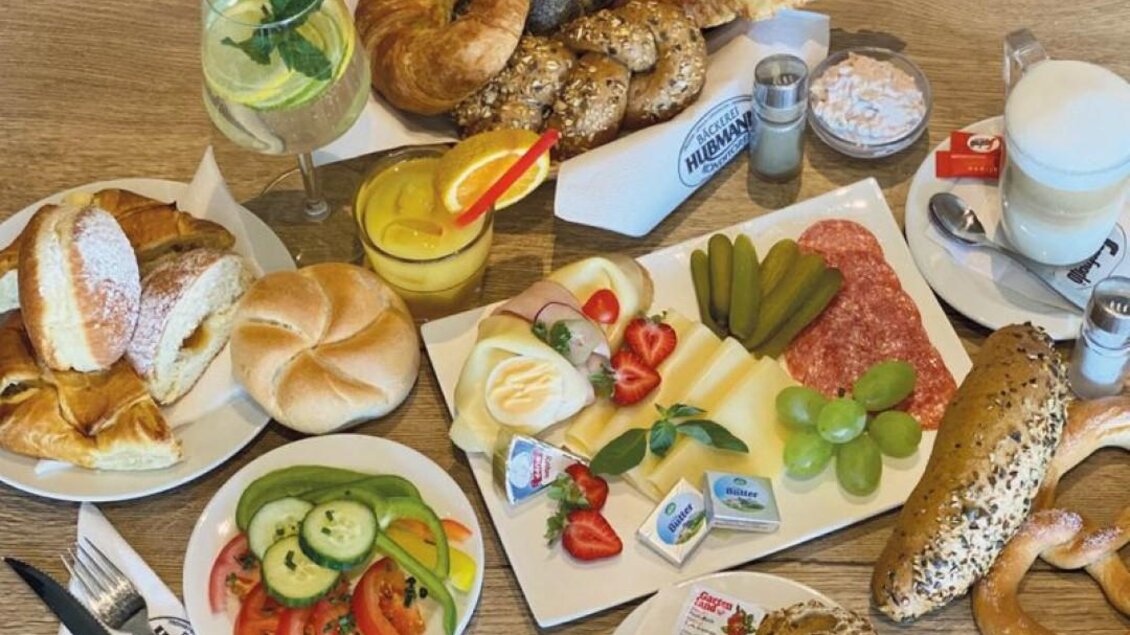 Ein täuschend attraktives Frühstücksarrangement mit verschiedenen Brötchen, Aufschnitt, Käse, frischem Obst und Gemüse. Dazu erfrischende Getränke serviert in bunten Gläsern. | © Bäckerei-Konditorei Hubmann Josef GmbH
