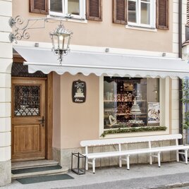 A charming shop with wooden windows and an inviting front door. The building has a pink facade and an awning. | © Café - Konditorei - Lebzelterei Ebner