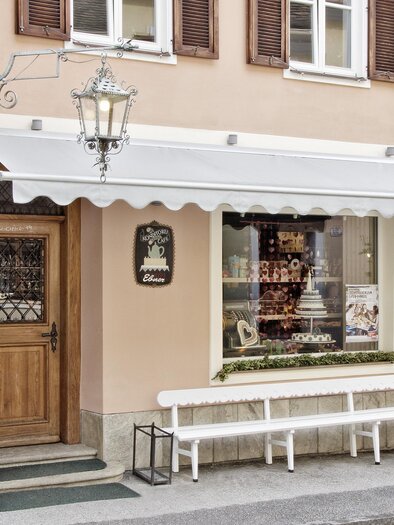 A charming shop with wooden windows and an inviting front door. The building has a pink facade and an awning. | © Café - Konditorei - Lebzelterei Ebner