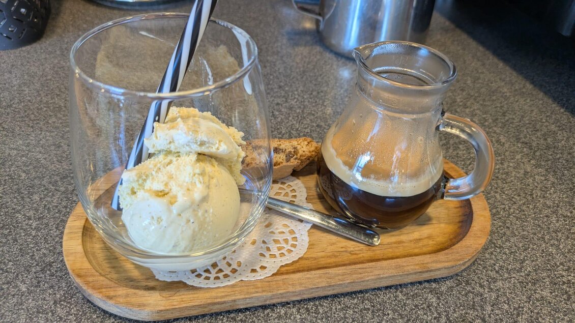 Ein Glas mit Eiscreme und zwei Strohhalmen steht auf einem Holzbrett. Daneben befindet sich ein kleines Gefäß mit einer Flüssigkeit und einer Kekse.