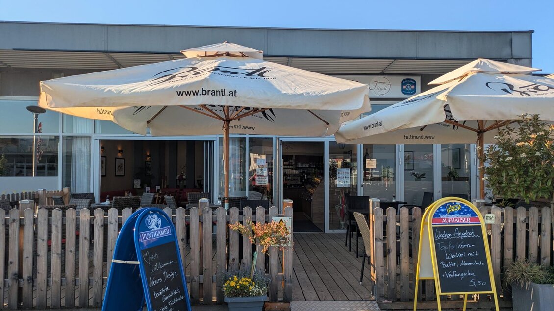 Ein gemütliches Restaurant mit Sonnenschirmen und einer Holzterrasse. Zwei spezielle Menütafeln stehen vor dem Eingang.