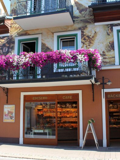 Ein einladendes Café mit einer Bäckerei im Erdgeschoss. Blühende Pflanzen schmücken den Balkon über der Eingangstür. | © TVB Ausseerland - Salzkammergut_Viola Lechner
