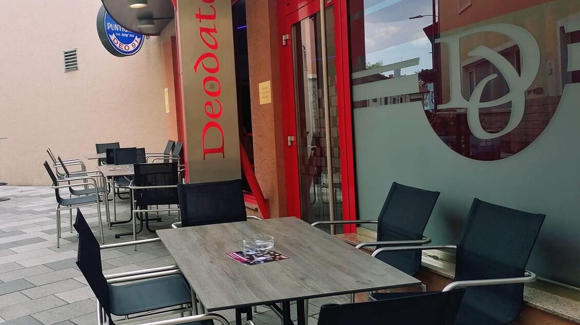 Ein gemütlicher Außenbereich eines Cafés mit schwarzen Stühlen und einem Tisch. Die Wände sind rot und es gibt große Fenster, die viel Licht hereinscheinen lassen. | © Susanne Seidl