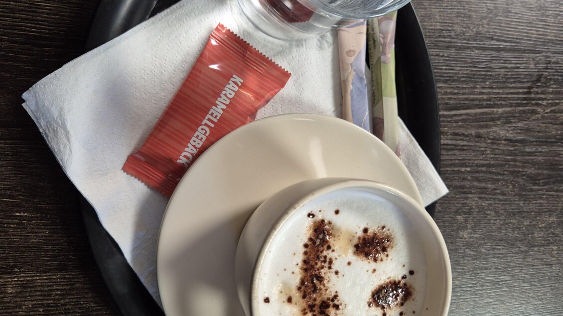 Eine Tasse Cappuccino mit Schaum und Schokolade auf einem Unterteller. Neben dem Kaffee stehen ein Glas Wasser und ein Päckchen Zucker. | © TV Murau