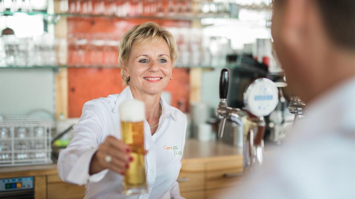 Eine lächelnde Bedienung reicht ein frisches Bier in einem modernen Restaurant. Im Hintergrund sind Gläser und eine einladende Atmosphäre zu sehen. | © Die Mosbacher´s