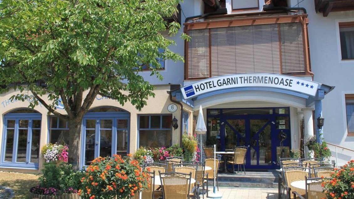 Ein einladendes Hotel mit der Beschriftung "Hotel Garni Thermeoase". Vor dem Gebäude befindet sich eine Terrasse mit Tischen und Stühlen, umgeben von Blumen. | © Café Hotel Garni Thermenaose
