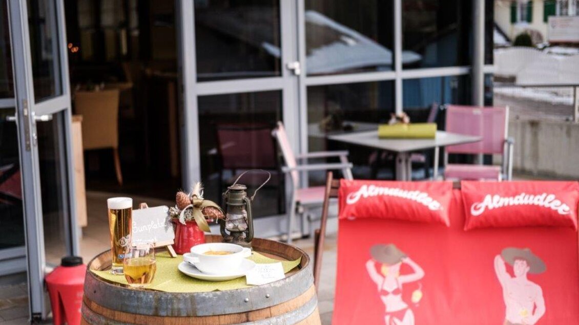 Eine gemütliche Terrasse mit einem Holztisch und einem Bierglas. Im Vordergrund steht eine dekorative Laterne und im Hintergrund sind Liegestühle mit bunten Aufdrucken. | © Café Oase