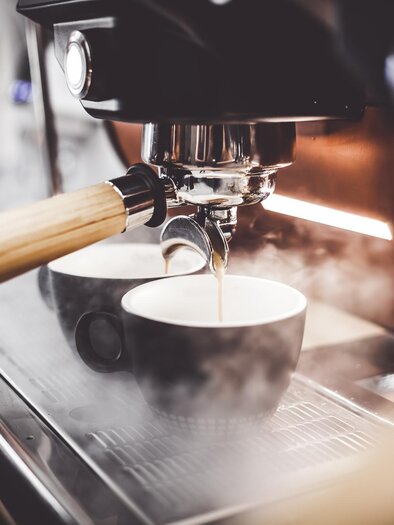 Eine Espresso-Maschine bereitet frischen Kaffee zu. Zwei schwarze Tassen stehen bereit, während Dampf aufsteigt. | © TV Südsteiermark - Adobe Stock