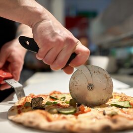 Ein Pizzabäcker schneidet eine köstliche Pizza mit einem Pizzaschneider. Die Pizza ist belegt mit frischem Gemüse und Käse. | © Adobe Stock_Pizzeria 2