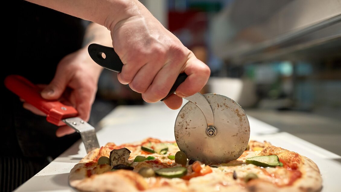 Ein Pizzabäcker schneidet eine köstliche Pizza mit einem Pizzaschneider. Die Pizza ist belegt mit frischem Gemüse und Käse. | © Adobe Stock_Pizzeria 2
