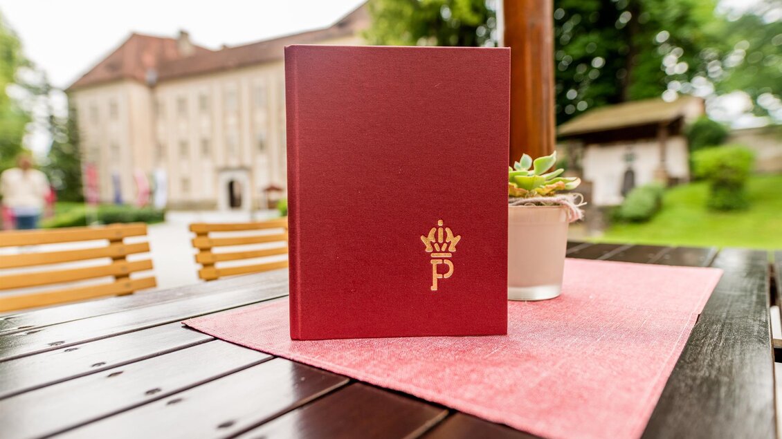 <p>Speisekarte auf Tisch im Café-Restaurant Piber mit Gastgarten und Gebäude im Hintergrund.</p> | © Lipizzanerheimat_Die Abbilderei