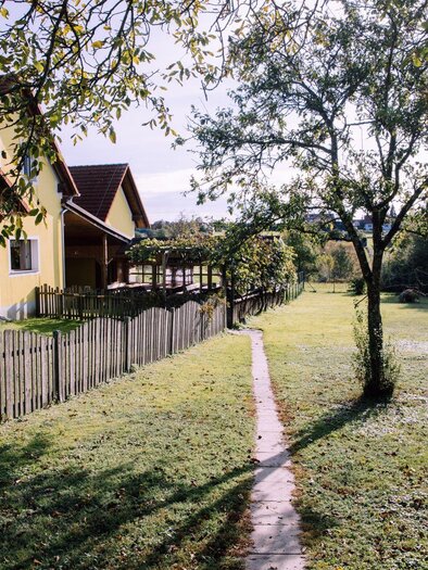 Ein gelbes Haus mit einem gepflegten Garten und einem Weg. Im Hintergrund ist ein Spielplatz mit Spielgeräten und ein großer Baum zu sehen. | © Kurkommission Bad Blumau