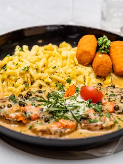 Ein köstliches Gericht mit Schnitzel in einer Sahnesauce, serviert mit Spätzle und Kroketten. Geprägt von frischem Gemüse und aromatischen Kräutern. | © Stephan Friesinger