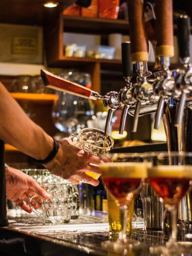 Ein Barkeeper zapft Bier in ein Glas. Im Vordergrund sind mehrere Getränke in verschiedenen Gläsern sichtbar.