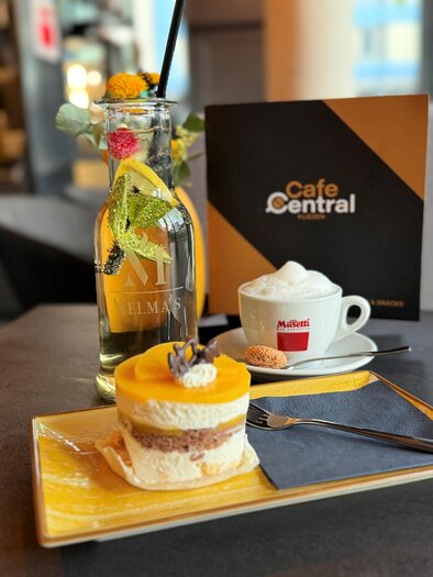 Ein leckerer Kuchen mit einer Mango-Deckschicht und einer Tasse Kaffee auf einem Tisch. Im Hintergrund steht eine Flasche und eine Speisekarte des Café Central. | © Cafe Central