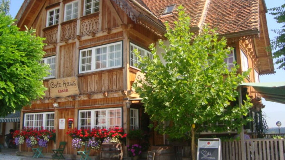 Ein charmantes, traditionelles Holzhaus mit blühenden Blumen und grünen Bäumen. Die Atmosphäre ist einladend und gemütlich. | © Cafehaus Ebner