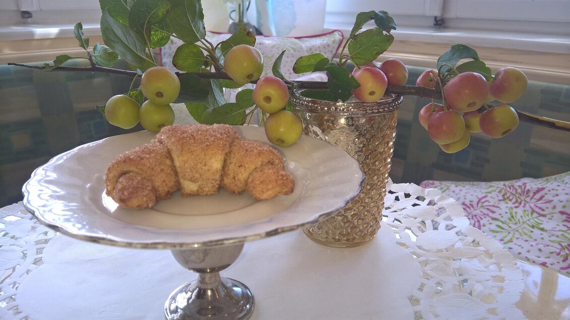 Ein Croissant auf einem silbernen Teller, umgeben von frischen Früchten und einer dekorativen Vase. Der Tisch ist mit einem feinen Tischläufer gedeckt. | © Cafehaus Ebner