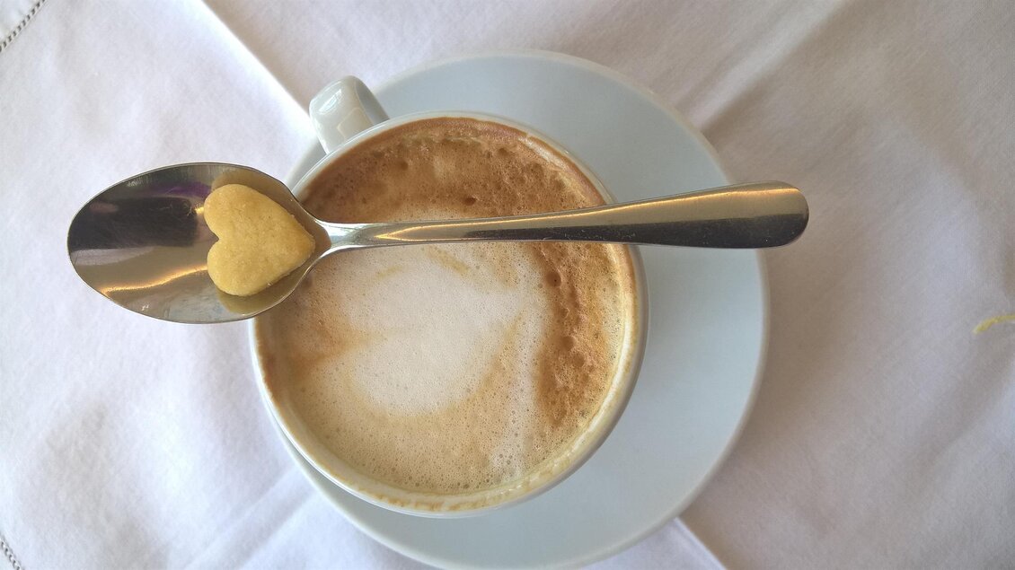 Eine Tasse Kaffee mit aufgeschäumter Milch und einem Löffel, auf dem ein Zuckerstück in Herzform liegt. Der Hintergrund ist hell und einfach gehalten. | © Cafehaus Ebner