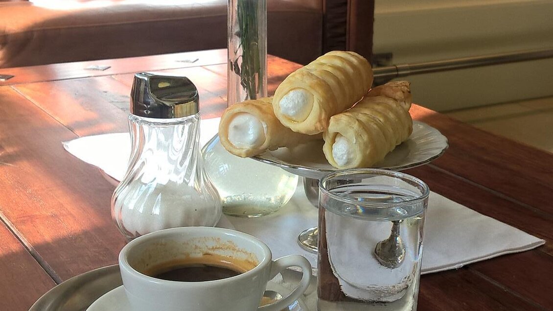 Ein gedeckter Tisch mit einer Tasse Kaffee, einem Glas Wasser und einem kleinen Behälter mit Milch. Dazu gibt es frische Backwaren und einen Blumenstrauß in einer Vase. | © Cafehaus Ebner