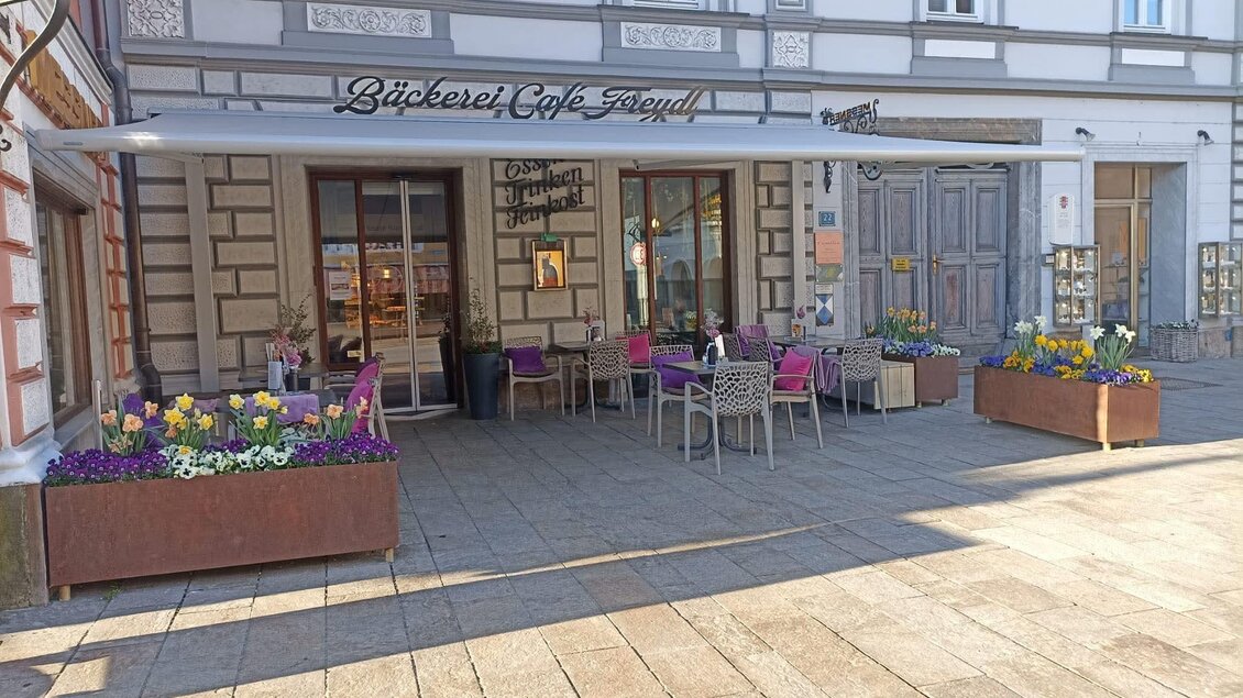 Ein gemütliches Café mit Außenterrasse und bunten Blumen in blühenden Kästen. Die Fassade des Gebäudes ist historisch gestaltet. | © Bäckerei Freydl