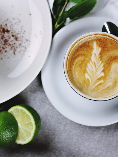 Ein schöner Kaffee mit kunstvollem Milchschaum und ein Stück Torte sind auf einem Tisch angerichtet. Frische Limetten und Minzblätter ergänzen das Bild. | © Pixabay
