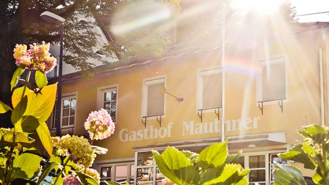 Ein Gasthof mit einem gelben Gebäude und Blumen im Vordergrund. Die Sonne strahlt hell und schafft eine einladende Atmosphäre. | © GH Mauthner Kirchenwirt