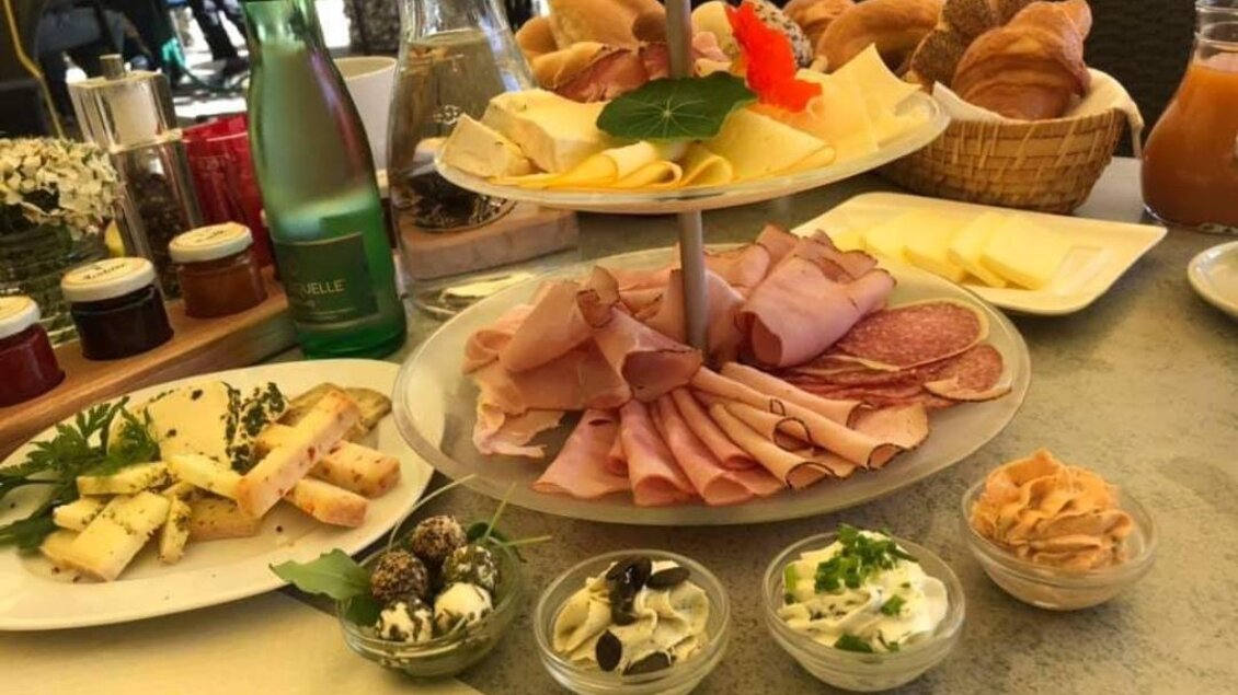 Ein reichhaltiges Frühstücksbuffet mit verschiedenen Käsesorten, Wurst und frischem Brot. Dazu sind kleine Schalen mit Aufstrichen und Marmelade angeordnet.