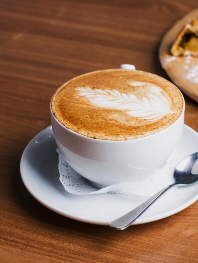 Eine Tasse Kaffee mit kunstvollem Milchschaum steht auf einem Unterteller. Im Hintergrund ist ein Stück Kuchen zu sehen. | © AdobeStock_171889547-Galina Zhigalova