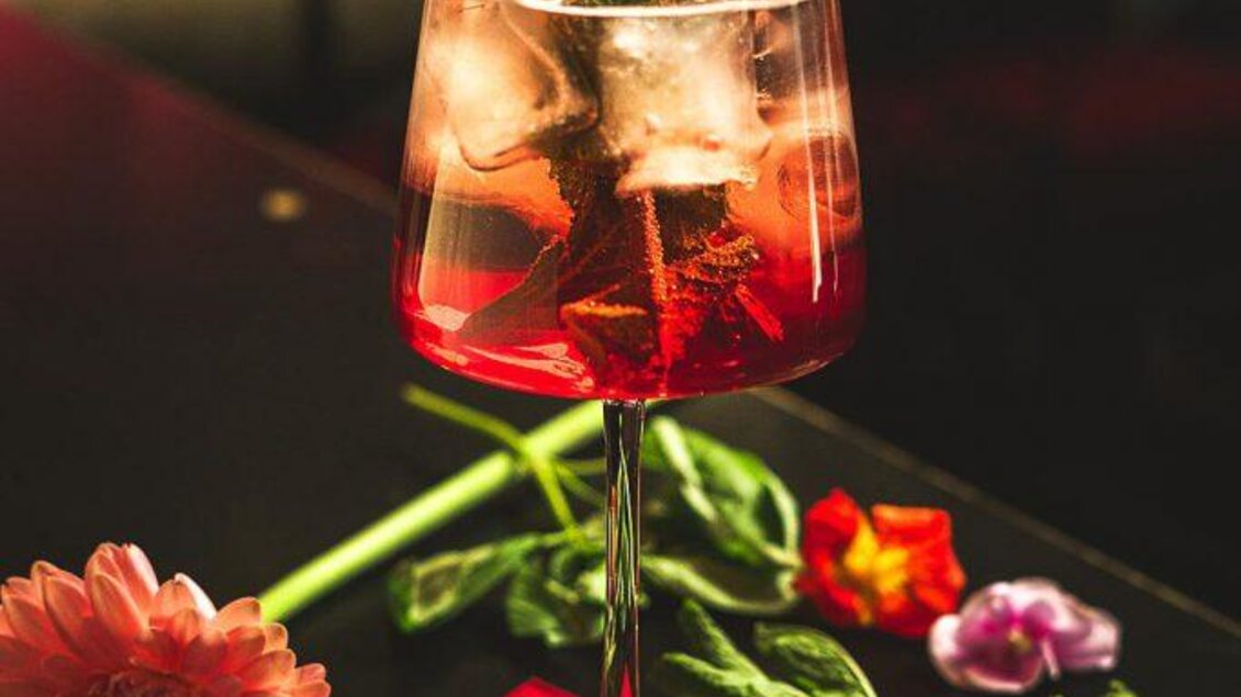 Ein farbenfroher Cocktail in einem hohen Glas mit frischen Minzblättern und Eiswürfeln. Um das Glas liegen bunte Blumen und grüne Blätter. | © Cafe Bar Maximilian