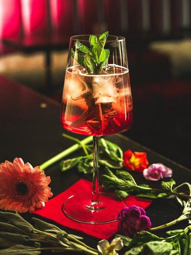 Ein farbenfroher Cocktail in einem hohen Glas mit frischen Minzblättern und Eiswürfeln. Um das Glas liegen bunte Blumen und grüne Blätter. | © Cafe Bar Maximilian