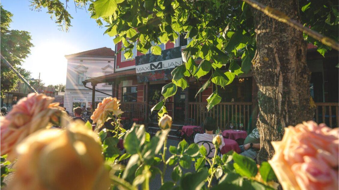 Ein gemütliches Restaurant umgeben von grünen Bäumen und blühenden Rosen. Die Sonne scheint sanft auf die Fassade des Gebäudes. | © Maximilian