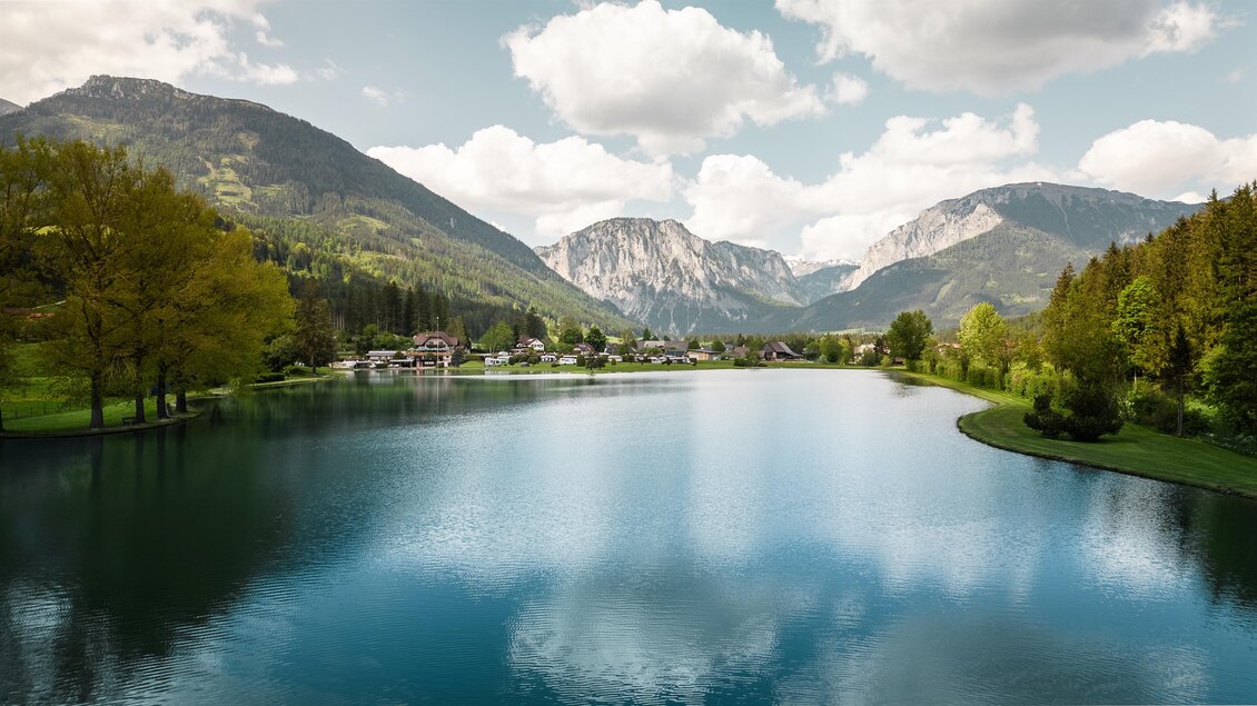 Ein ruhiger See umgeben von grünen Bergen und Bäumen. Der Himmel ist teilweise bewölkt und spiegelt sich im Wasser. | © Hannes Zenz
