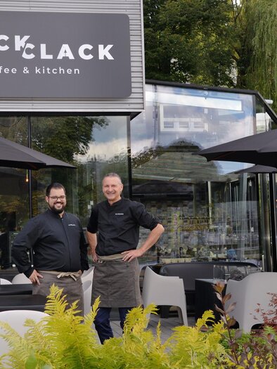 Two men are standing in front of a modern café named "CLACK coffee & kitchen." The outdoor area is furnished with tables, chairs, and umbrellas. | © Click Clack - Silvia Lambauer Fotografie