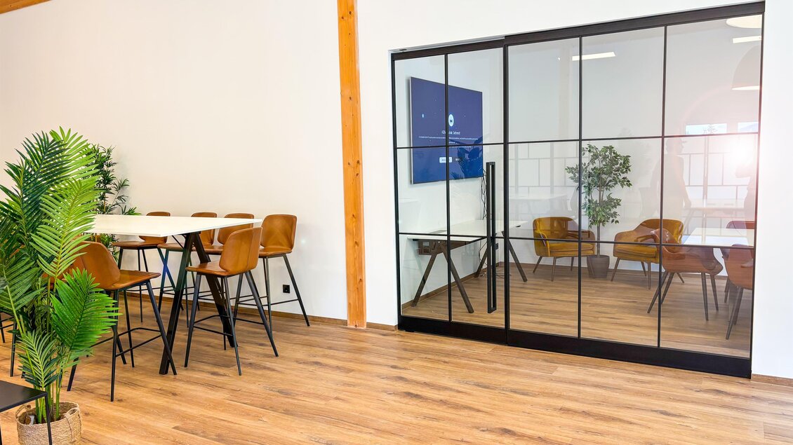 Ein moderner Büroraum mit einem Tisch, Stühlen und Pflanzen. An einer Wand befindet sich eine große Glastür, die einen weiteren Bereich zeigt. | © Coworking Space