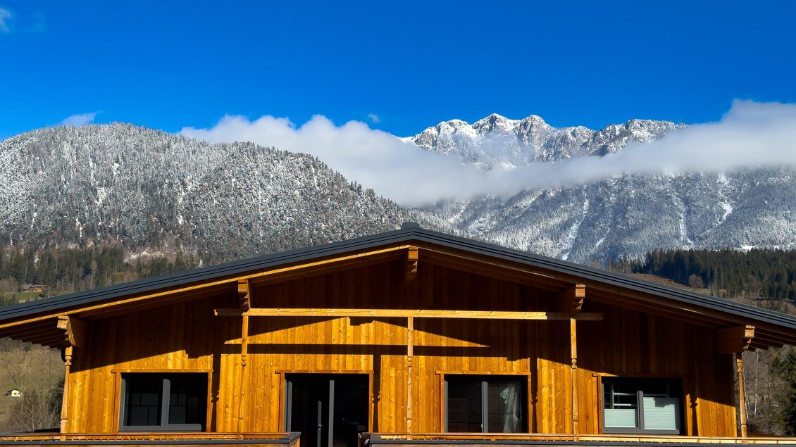 Ein modernes Holzhaus mit einem Balkon steht vor schneebedeckten Bergen. Der Himmel ist klar und blau, was eine schöne Landschaft schafft. | © Coworking Space