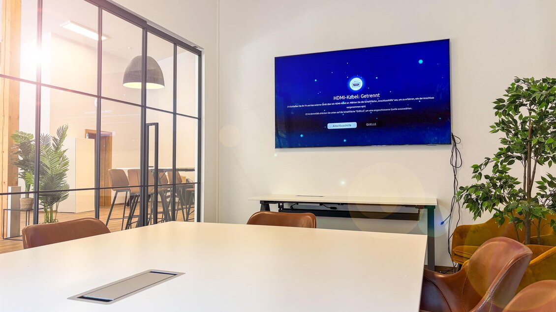 Ein modernes Konferenzzimmer mit einem großen Tisch und stilvollen Stühlen. An der Wand hängt ein Bildschirm, der Informationen anzeigt. | © Coworking Space