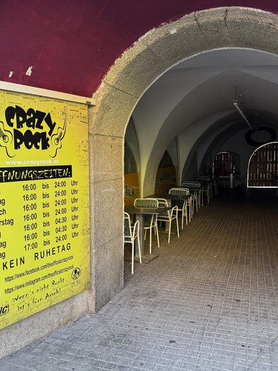 An unconventional entrance to a venue named "Crazy Pot". Opening hours are posted on the walls, and some tables and chairs can be seen. | © TV Hochsteiermark