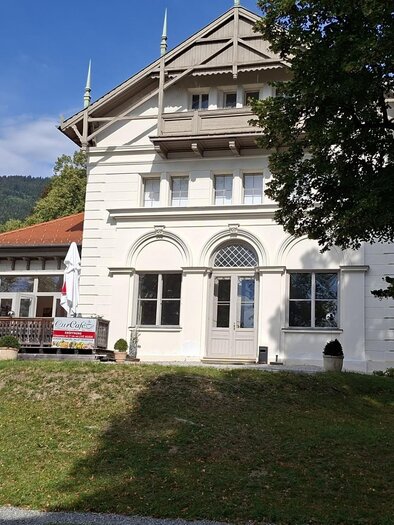 White building of the CurCafés in St. Radegund with terrace, trees, and wooded hill in the background. | © Bernhard Glauninger