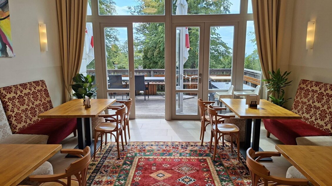 Innenraum des CurCafés mit Holztischen, Sitzplätzen, Teppich und Blick auf die Terrasse mit Bäumen im Hintergrund. | © Bernd Glauninger