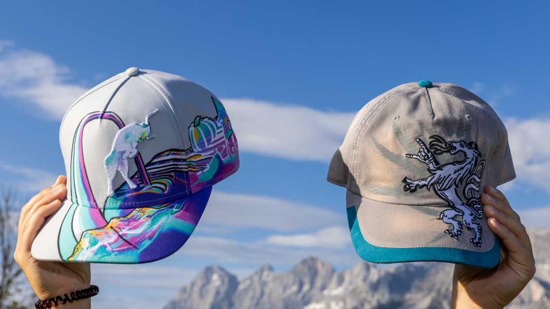 Zwei stylische Caps werden in die Kamera gehalten, vor einer malerischen Berglandschaft. Der Himmel ist klar und blau, was das Bild lebendig macht.