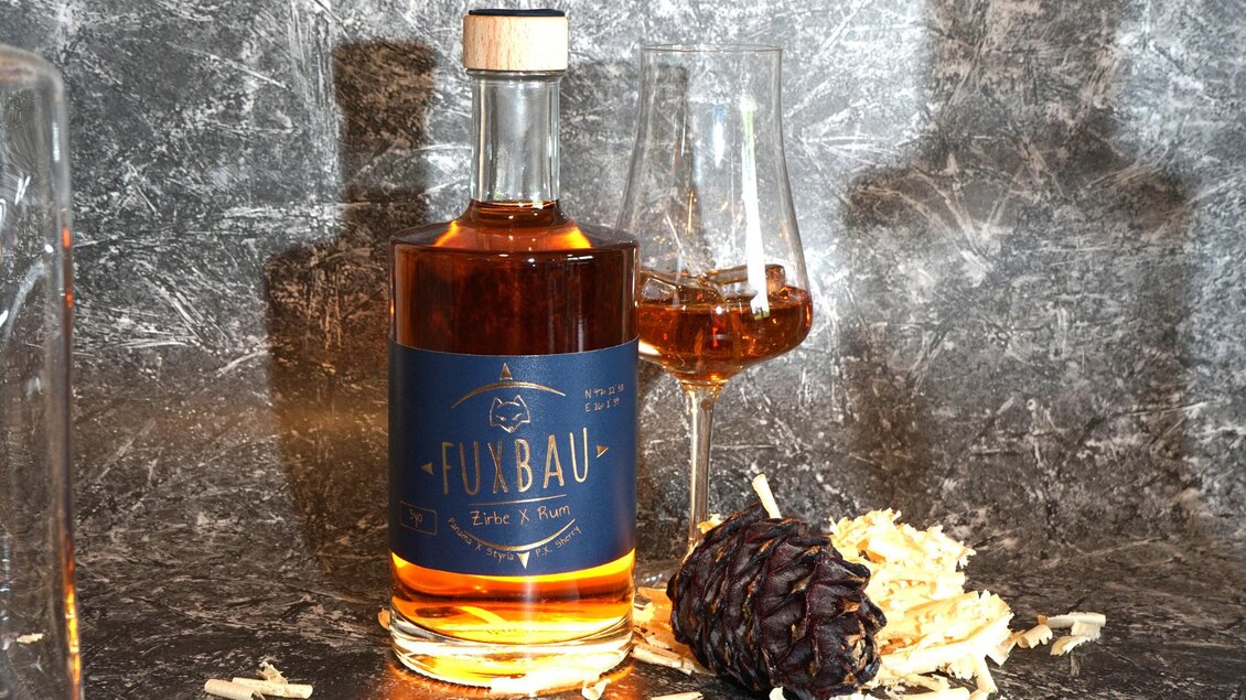 Eine Flasche mit dem Namen "Fuxbau" steht neben einem Glas, gefüllt mit einem bernsteinfarbenen Getränk. Neben der Flasche liegen einige Holzspäne und eine Tannenzapfen.