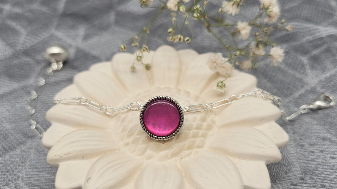 Ein elegantes Armband mit einem pinken Edelstein liegt auf einer blütenförmigen Unterlage. Im Hintergrund sind zarte Blumen zu sehen.
