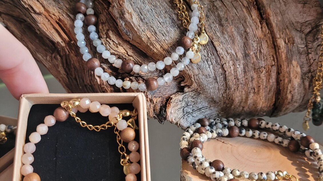 Schöne handgefertigte Halsketten und Armbänder aus verschiedenen Steinperlen liegen auf einem Holzstück. Eine elegante Verpackung für ein Armband wird von einer Hand gehalten.