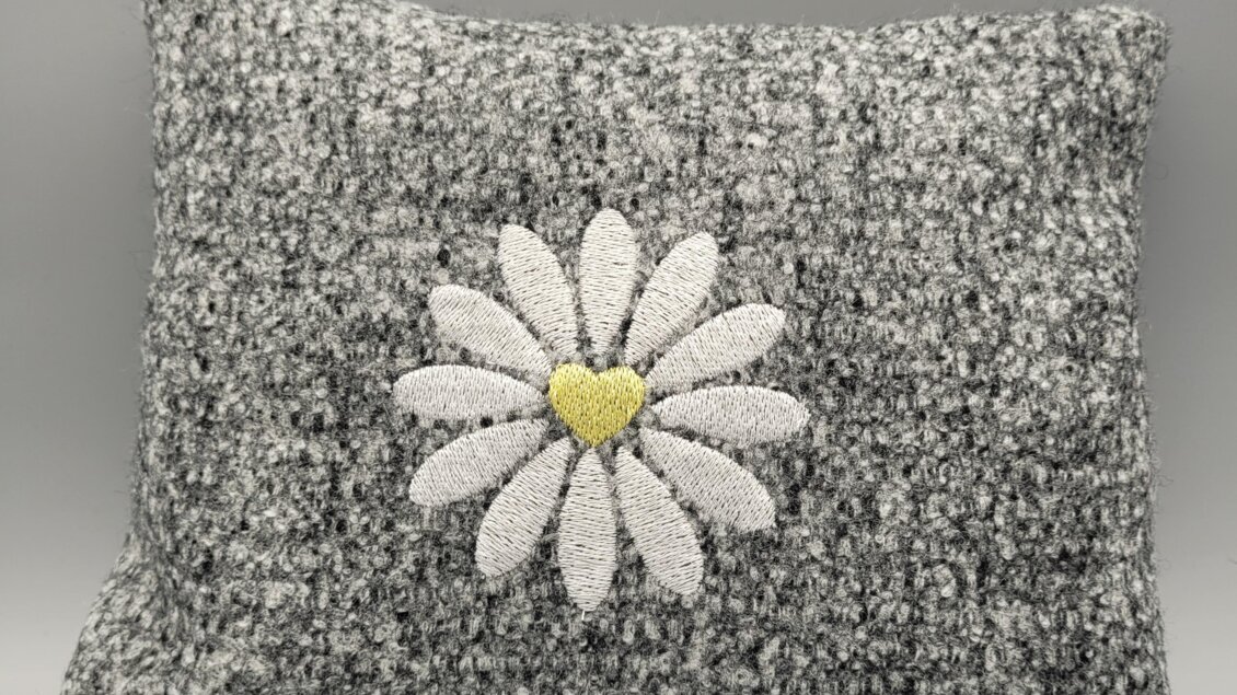 Ein graues Kissen mit einer weißen Blume und einem gelben Herzen in der Mitte. Das Design ist schlicht und dennoch ansprechend.