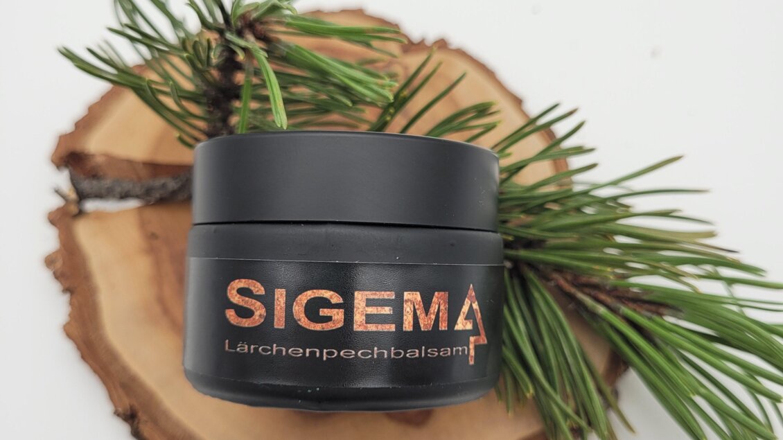 Ein schwarzes Cremeglas mit der Aufschrift "SIGEMA Lärchenpechbalsam" liegt auf einer Holzscheibe. Neben dem Glas sind einige Fichtenzweige angeordnet.