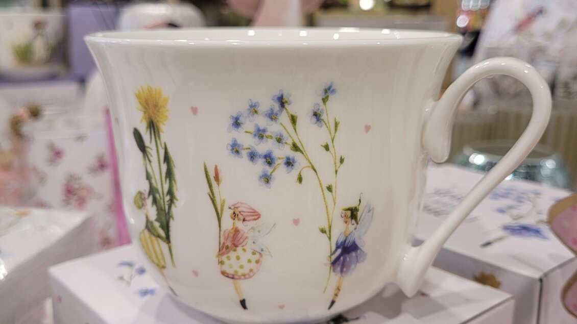 Eine dekorative Tasse mit Blumenmotiven und kleinen Figuren. Sie steht auf einer Box in einem geschmackvollen Geschäft.