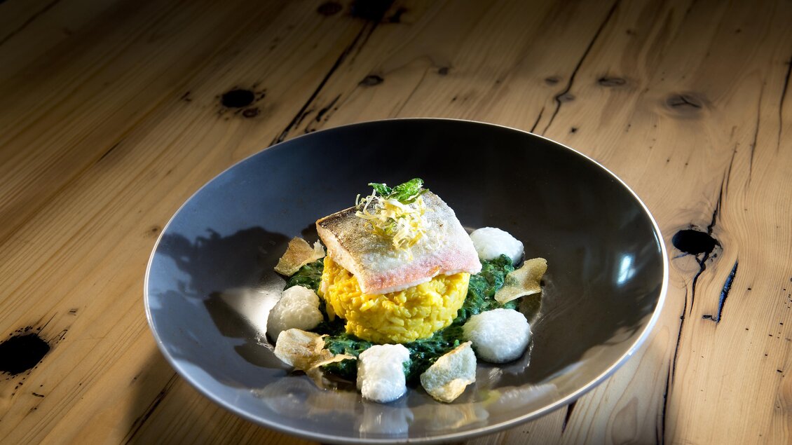 Ein kunstvoll angerichteter Teller mit Risotto, Fisch und grünem Püree. Dazu liegen einige knusprige Chips und kleine Beilagen auf dem Teller. | © A.M.J. Hotel KG
